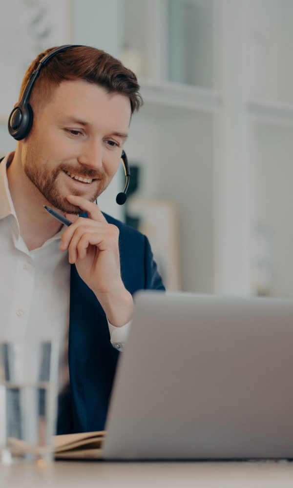 Professional male operator works in callcenter uses headset and laptop computer talks on video call or virtual webcam event wears formal clothes poses against cozy interior has online meeting